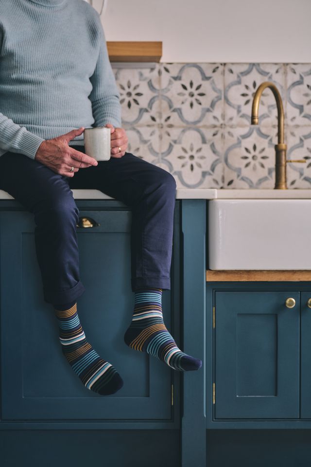 A person sitting on a blue kitchen cabinet holds a white mug, wearing navy trousers and striped crew socks in blue, brown, and cream, with a patterned tile backsplash and brass faucet behind them.