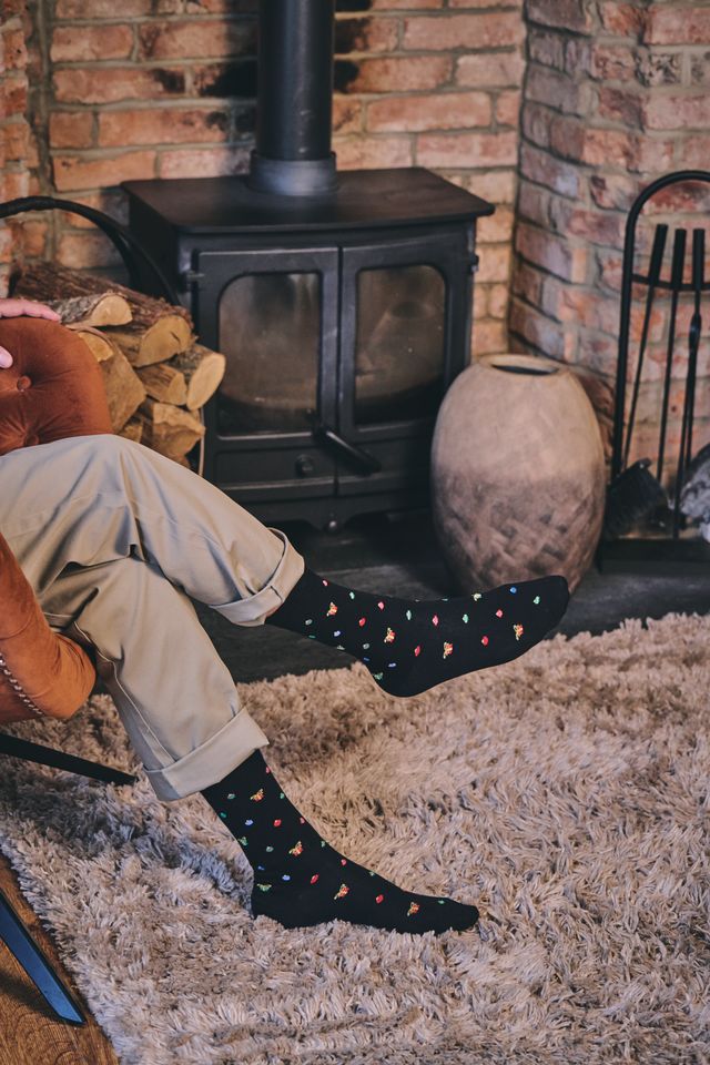 A person sitting in a chair with legs crossed, wearing black crew socks with a small multicoloured pattern. The setting is a cosy room with a brick fireplace, a black wood-burning stove, a shag rug, and a large ceramic pot.