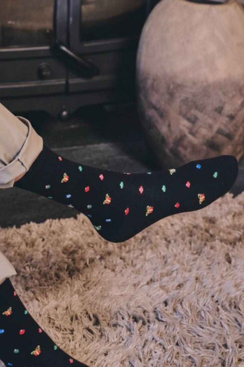 Black crew socks with a colourful pattern worn on a person's foot in a cosy indoor setting with a shag rug and fireplace visible.