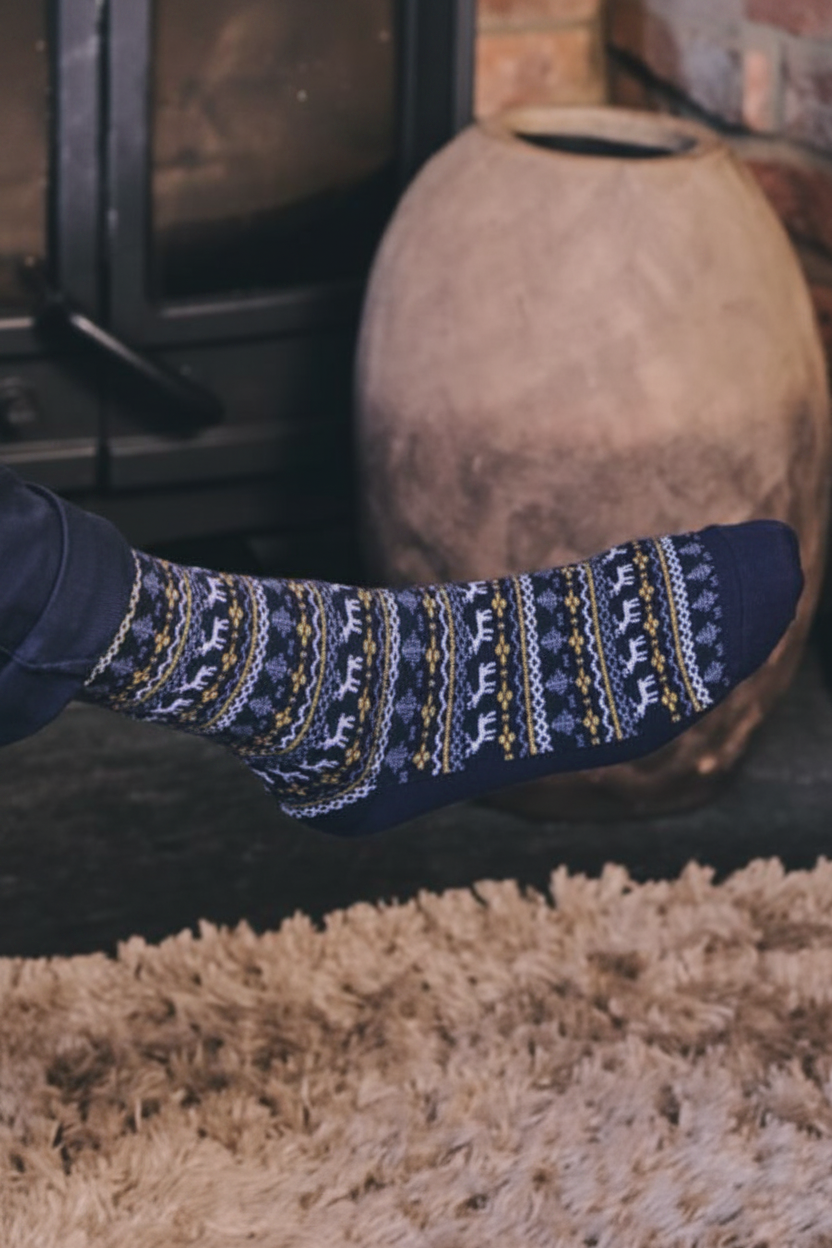 A close-up of a person's foot wearing a navy blue knit sock with a traditional Nordic fair isle pattern in white, yellow, and gray, set against a cozy indoor background with a fireplace and a large ceramic pot.