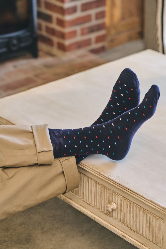 A person wearing navy crew socks with small multicoloured polka dots, paired with beige trousers, with their legs stretched across a light-colored wooden bench in a cosy indoor setting.