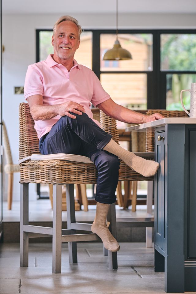 A smiling man with gray hair sits in a wicker chair at a kitchen island, wearing a pink polo shirt, dark trousers, and beige socks in a bright modern kitchen.