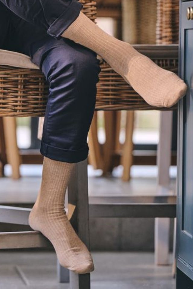 A man sitting at a wicker bar stool in a cafe, wearing a pink polo shirt, dark trousers, and beige crew socks.