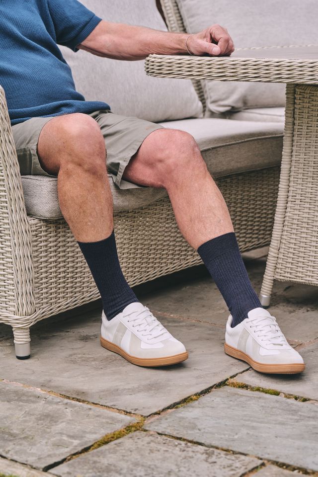 A person seated in a wicker patio chair wearing dark navy crew socks and white low-top sneakers with gum soles, with a blue shirt and khaki shorts visible.