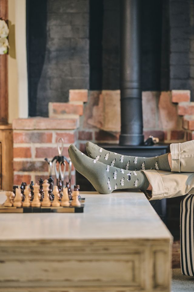 A person's legs and feet resting on a table in a cozy room, wearing gray knit socks with a white pattern. A chessboard sits on the table in the foreground, with a brick fireplace and dark wood stove in the background.