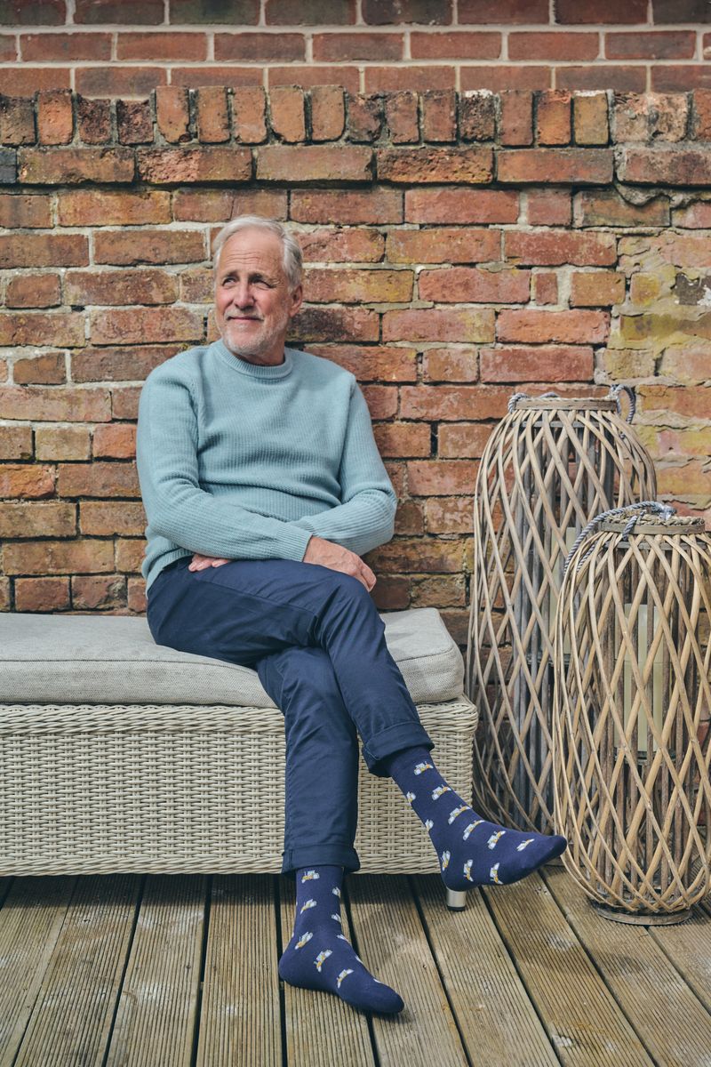 A man sits on a wicker patio bench against a red brick wall, wearing a light blue sweater, navy trousers, and navy socks. Two large woven decorative baskets are on a wooden deck beside him.