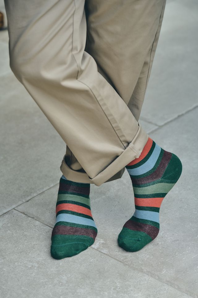 A close-up of a person's legs and feet wearing cuffed khaki trousers and a pair of multicoloured striped socks with green, red, orange, and blue bands, standing on a grey concrete surface.