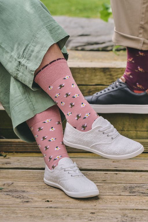 Pink crew socks with a black and white butterfly pattern, worn by a person sitting on wooden steps outdoors. The person is wearing a sage green skirt and white sneakers, with another person's legs visible in the background wearing khaki pants and dark shoes.