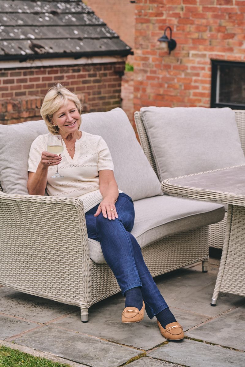 A woman sits smiling in a wicker patio lounge chair with a glass of white wine, set in an outdoor patio area with brick walls and a matching wicker table.