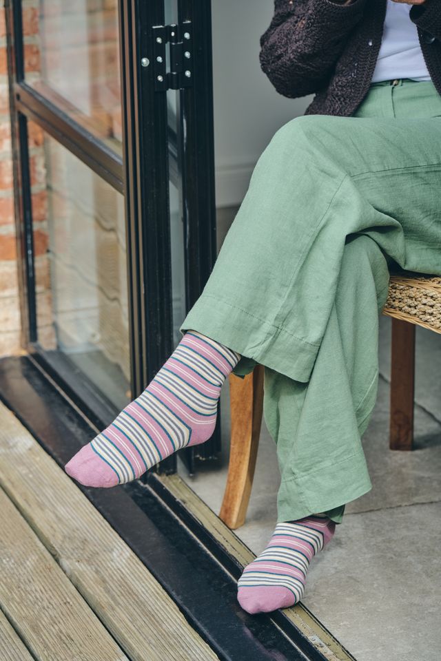 A person seated on a chair by a glass door wearing pink striped socks with thin multicolor lines, sage green wide-leg pants, a dark knit cardigan, and a white top.