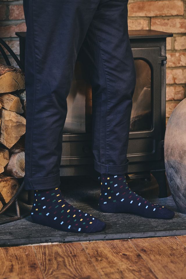 A person wearing dark navy socks with small multicolored diamond patterns, paired with dark pants, standing in front of a black stove and brick fireplace.