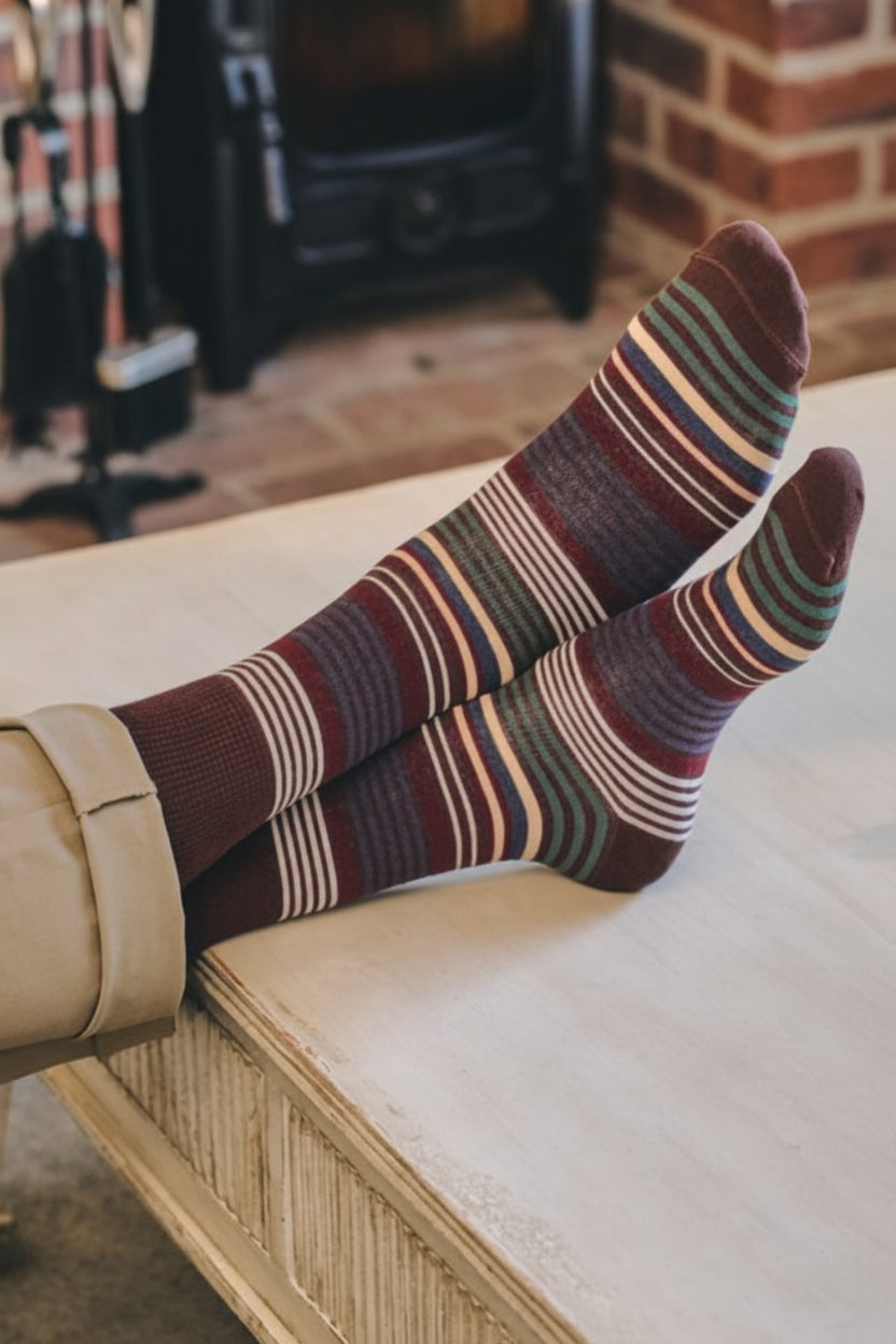 A person's legs crossed over a light wood bench, wearing striped knit socks in shades of burgundy, brown, beige, and navy, with a cozy brick fireplace and wood stove in the background.