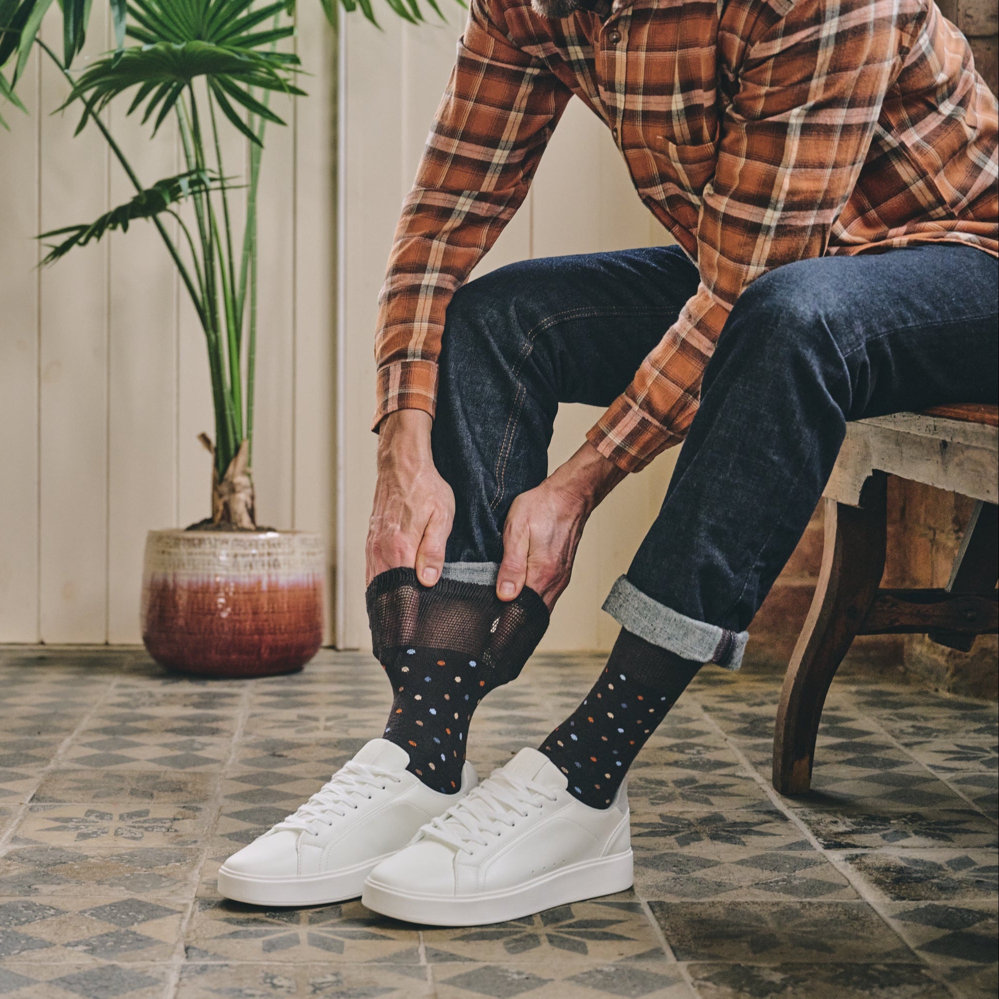 Person wearing plaid shirt, dark jeans, and white sneakers with a plant in the background.