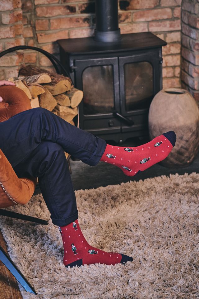 A person sitting in a chair with their legs crossed, wearing red crew socks with a small black and white pattern and dark navy pants, set in a cozy room with a brick fireplace, wood stove, and shag rug.