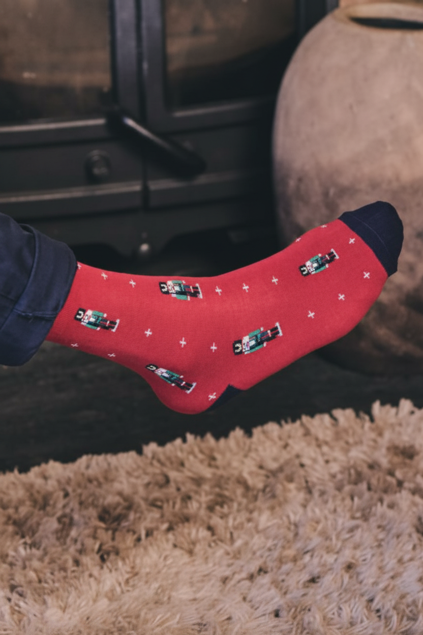 A person's foot wearing a red crew sock with a pattern of small black and white cars and white stars, paired with dark blue jeans, in a cosy indoor setting with a shag rug and fireplace.