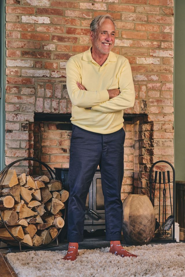 A smiling man with gray hair stands in front of a brick fireplace wearing a pale yellow long-sleeve polo shirt, navy pants, and brown leather shoes. A wood stack is visible on the left, and the background features a rustic brick fireplace and a shag rug.