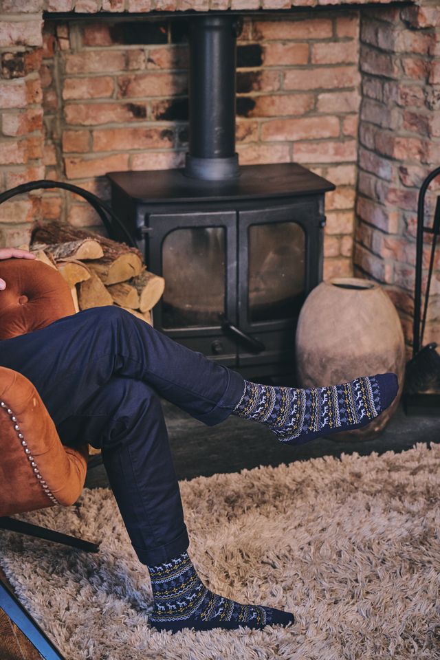 A person sitting in a brown armchair by a brick fireplace and wood-burning stove, wearing navy blue trousers and patterned knit socks in blue, white, and gray.
