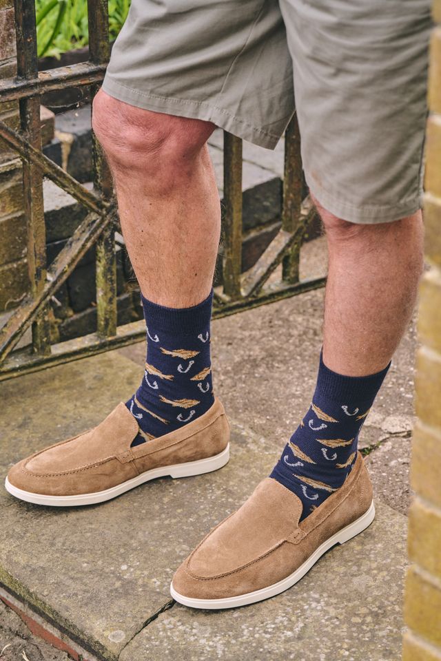 A person wearing navy blue crew socks with a repeating gold and white fish pattern, paired with tan suede slip-on shoes and khaki shorts, standing on a stone surface.