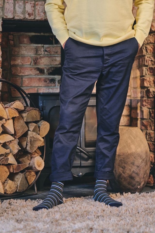 A person standing in front of a rustic brick fireplace wearing navy blue trousers, a pale yellow sweatshirt, and striped socks, with a stack of firewood and a shag rug visible.