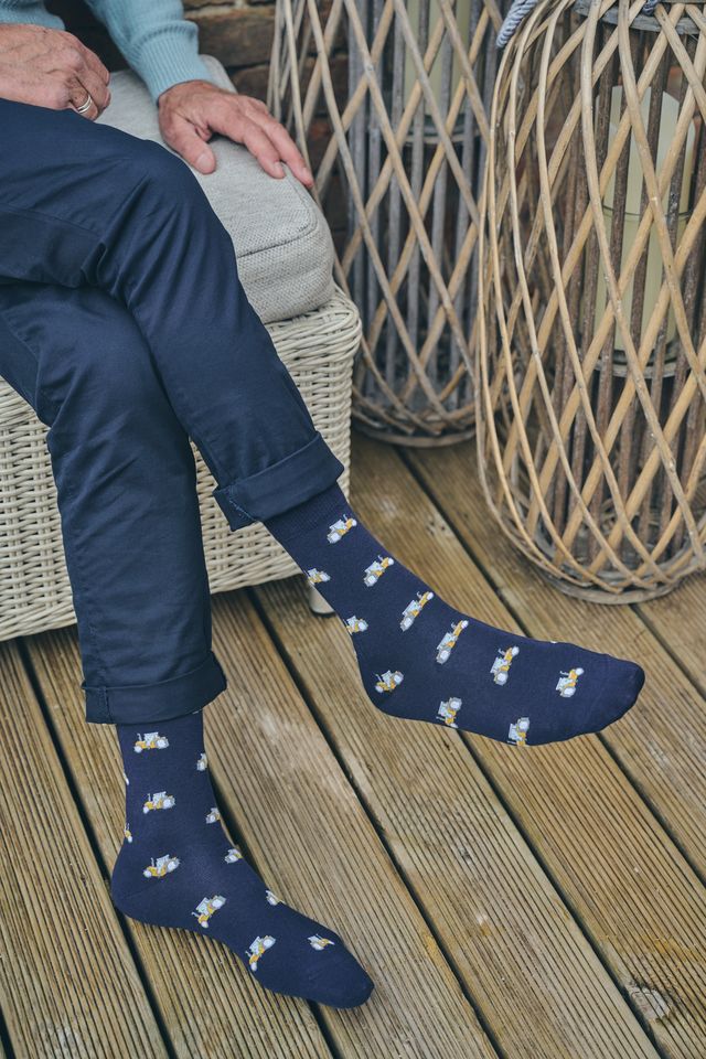 A person sitting on a wicker chair on a wooden deck wearing navy blue crew socks, paired with navy trousers.