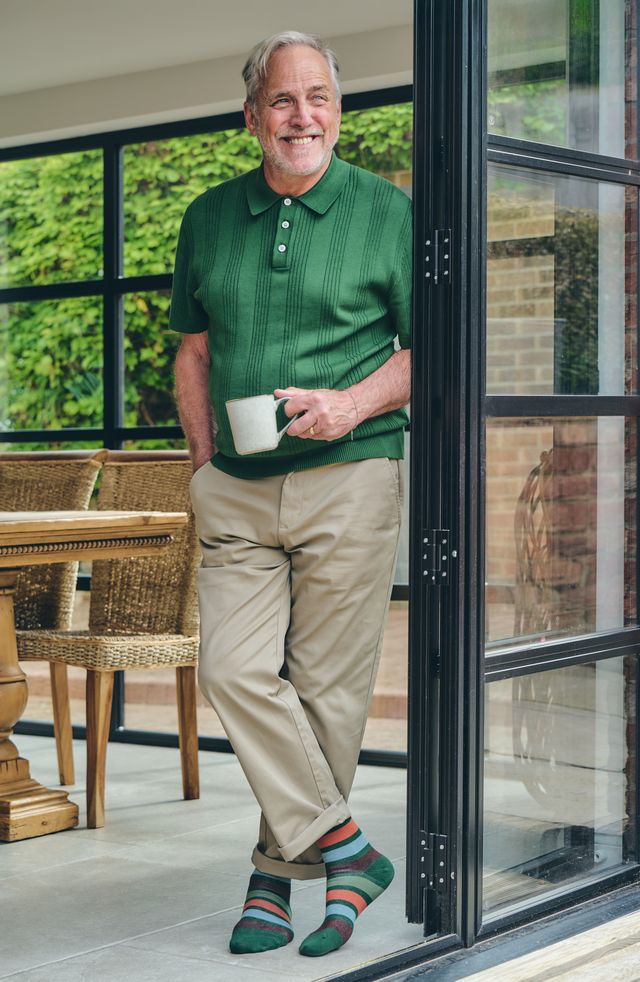 An older man stands in a doorway holding a white mug, wearing a green short-sleeve polo shirt, khaki trousers, and striped socks. The setting appears to be a modern home patio with large black-framed glass doors and outdoor greenery in the background.