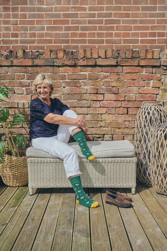 A woman sits on a wicker bench on a wooden deck in front of a brick wall, wearing green patterned socks with yellow toes and heels, white trousers, and a navy top. A pair of brown shoes sits on the deck beside her.