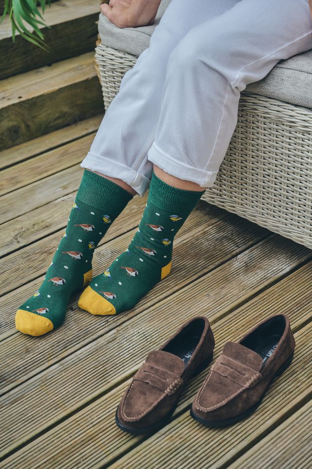 A person sitting on a wicker bench wearing dark green patterned socks with yellow toes and heels, paired with brown suede loafers on a wooden deck.