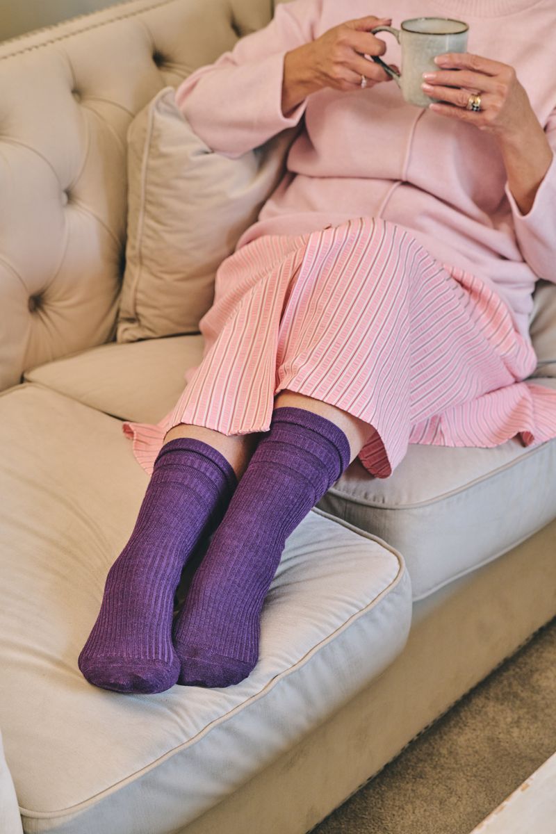 A person reclining on a cream tufted sofa wearing pink ribbed knit pants and purple ribbed knit socks, holding a light blue mug.