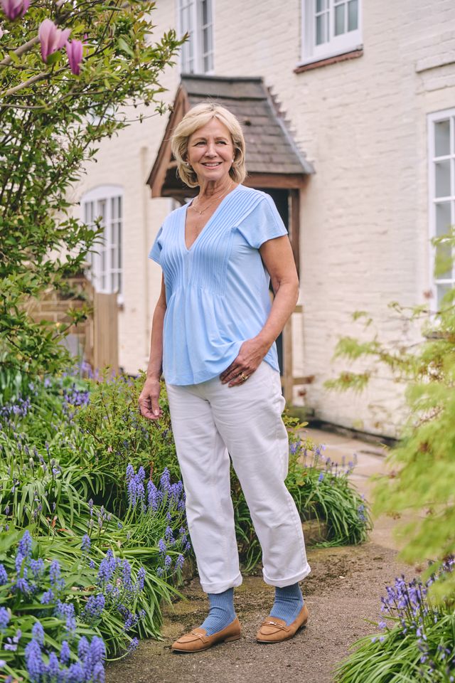 A smiling woman stands in a garden wearing a light blue short-sleeve V-neck top, white trousers, blue socks, and tan loafers, with a white brick house in the background.