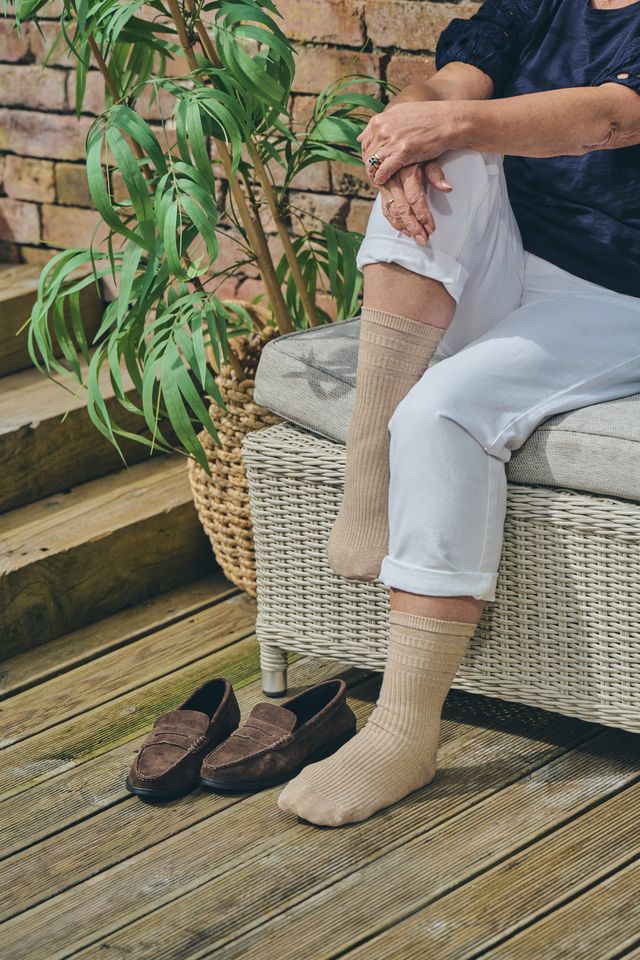 A person sitting on a wicker bench wearing light beige ribbed knee-high socks, with matching socks on the other foot and a pair of brown loafers on a wooden deck beside a potted plant.