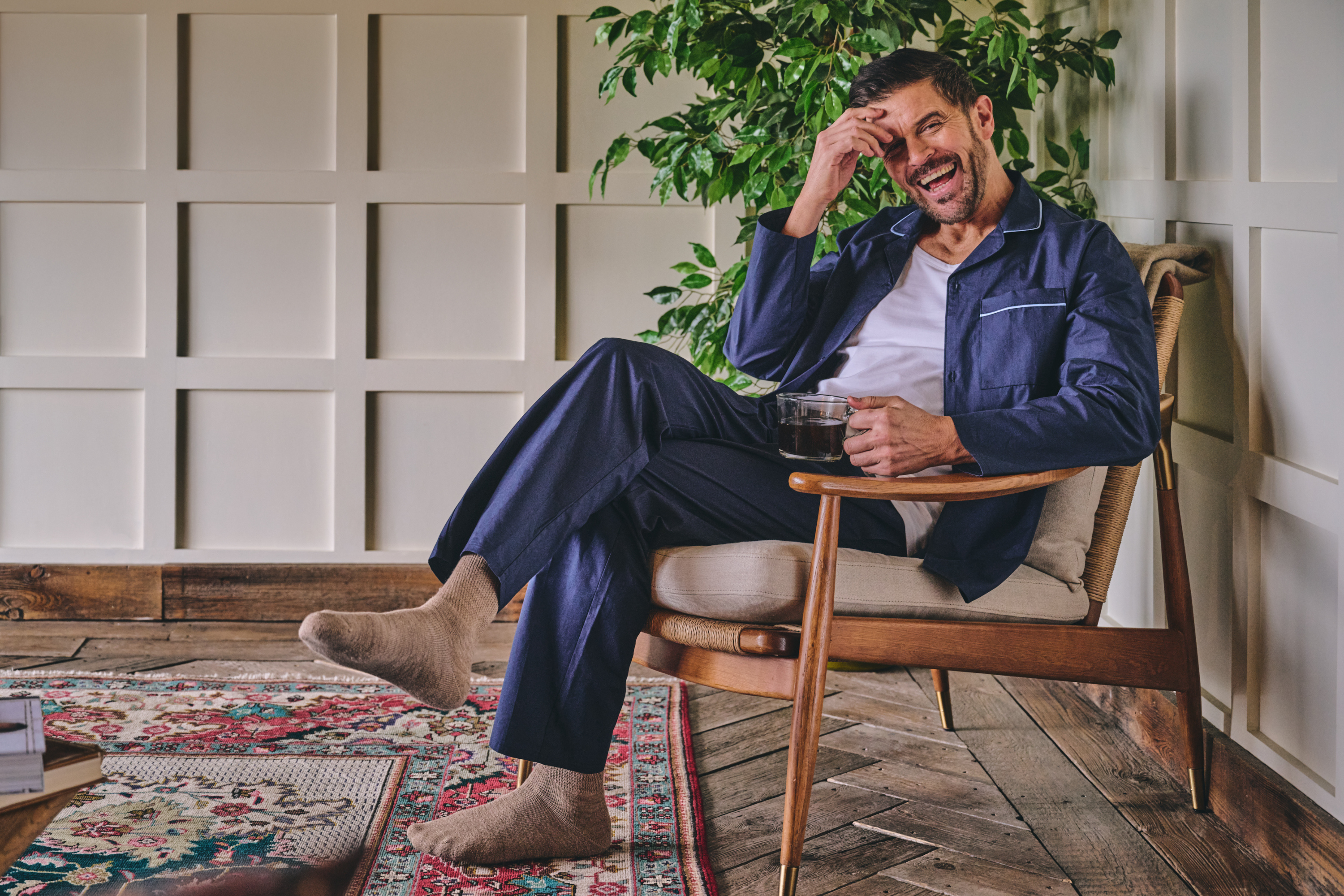 Man sitting in a chair in pyjamas and socks, holding a coffee, surrounded by plants and wooden flooring.