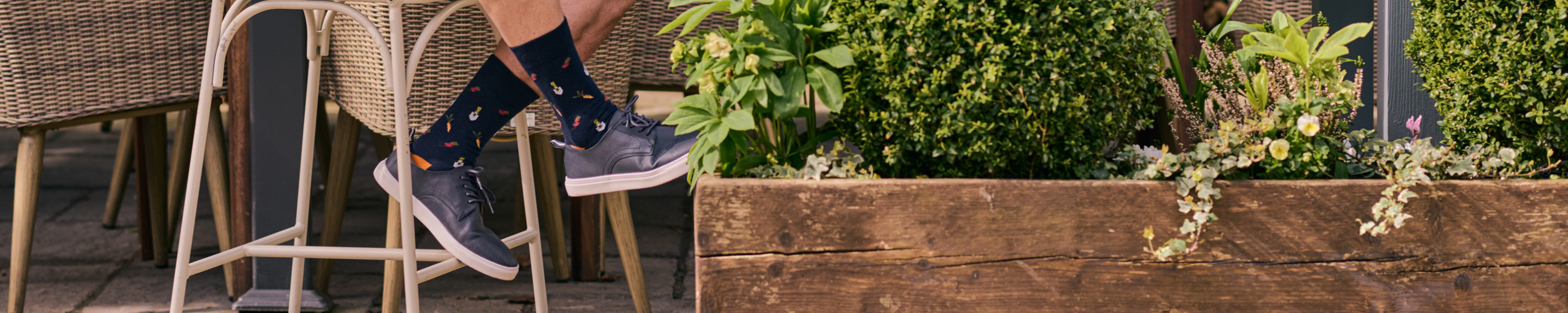 Person wearing blue sneakers and patterned socks sitting on a chair with a garden and wooden planter in the background.