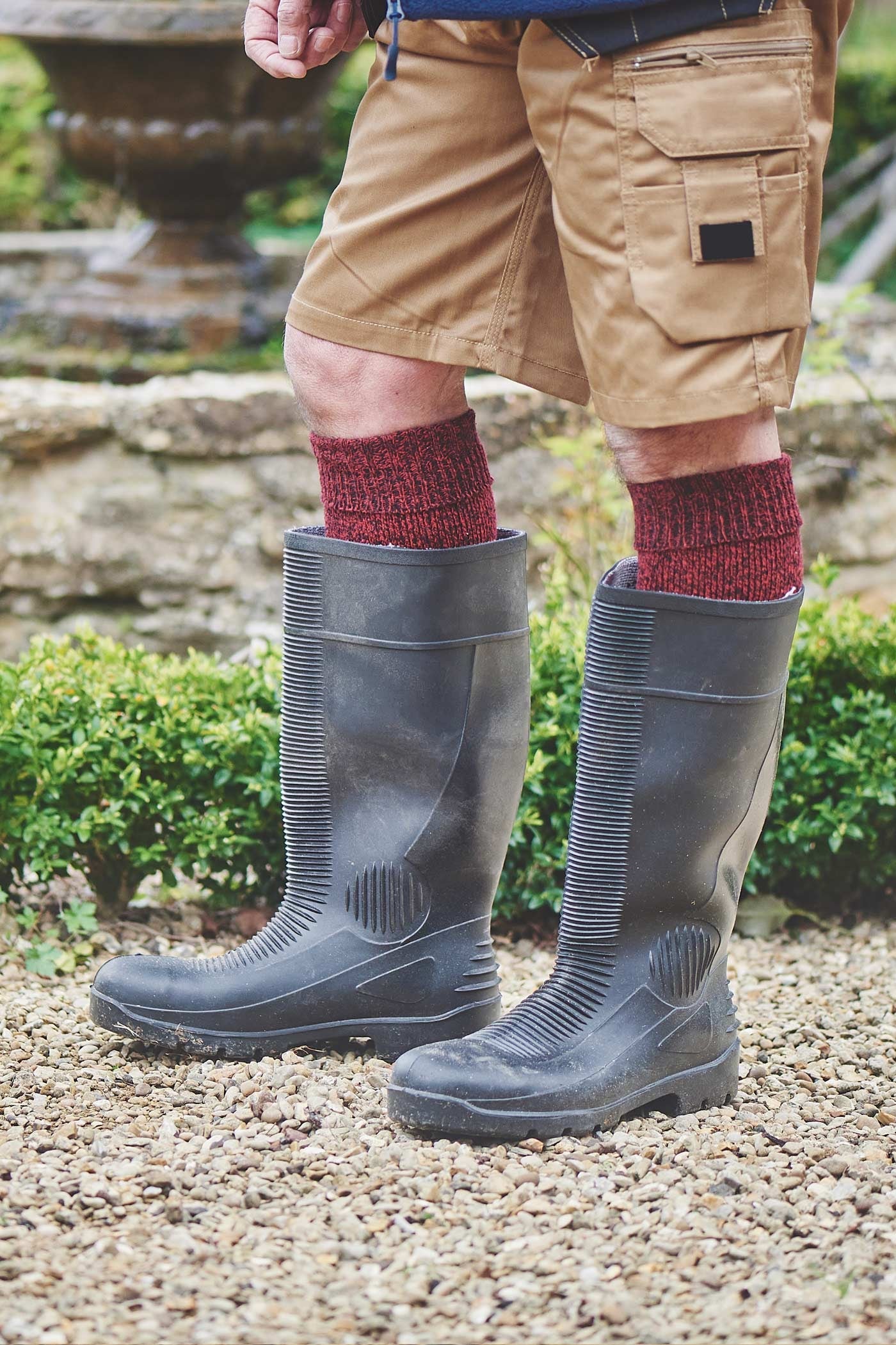 Person wearing brown cargo shorts, gray rubber boots, and red socks standing on a gravel path with greenery in the background.